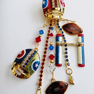 May include: A gold charm with a football helmet, football, and goal post design. The charm is decorated with red, white, and blue glitter and rhinestones. The charm is attached to a gold chain with red, white, and blue beads.