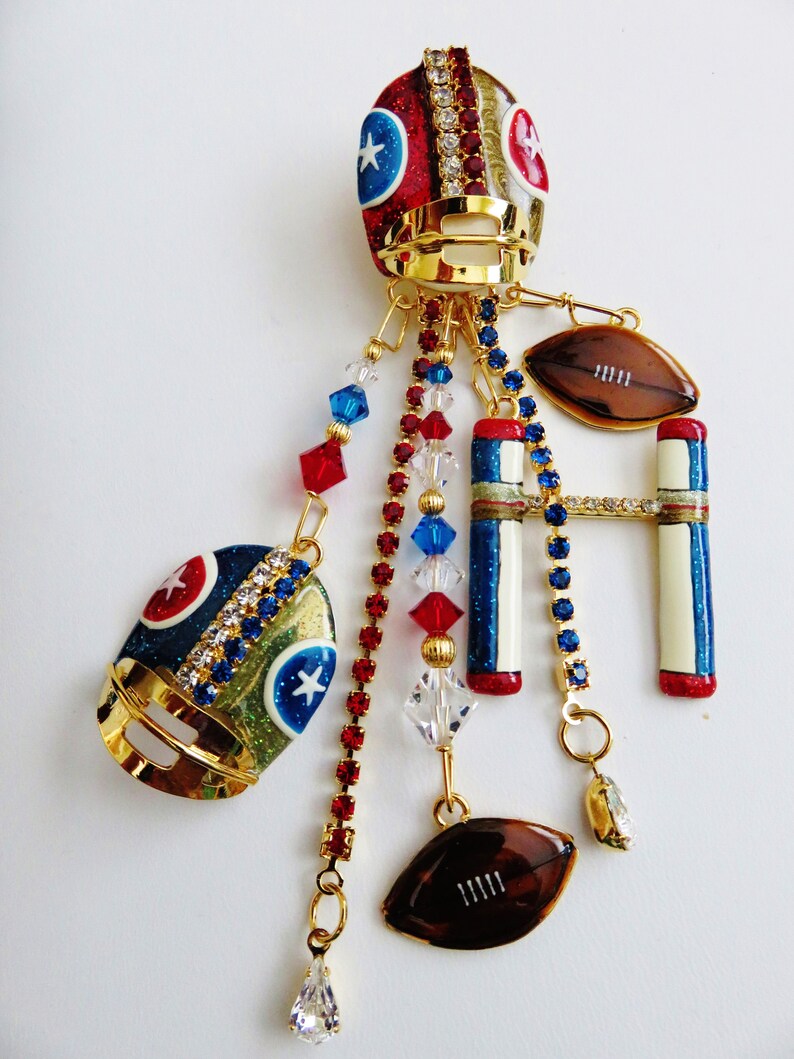 May include: A gold charm with a football helmet, football, and goal post design. The charm is decorated with red, white, and blue glitter and rhinestones. The charm is attached to a gold chain with red, white, and blue beads.