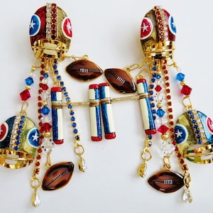 May include: A pair of gold football helmet charms with red, white, and blue details. Each helmet is adorned with rhinestones and has a chain with charms including footballs, stripes, and crystals.
