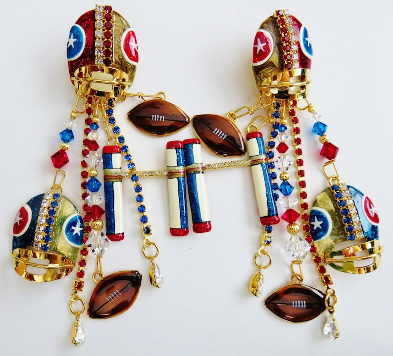 May include: A pair of gold football helmet charms with red, white, and blue details. Each helmet is adorned with rhinestones and has a chain with charms including footballs, stripes, and crystals.