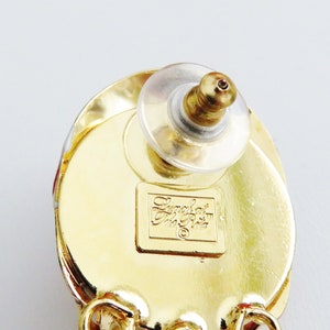 May include: Gold tone earrings with a red and white design. The back of the earring has a clear plastic post and a small gold label that reads "Emerald  Jewelry  Co."