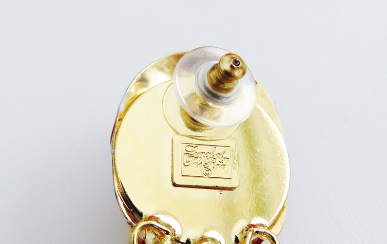 May include: Gold tone earrings with a red and white design. The back of the earring has a clear plastic post and a small gold label that reads "Emerald  Jewelry  Co."