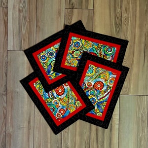 May include: A set of four square coasters with a vibrant floral pattern. Each coaster has a red border, a black patterned frame, and a colorful design featuring flowers and abstract shapes. The coasters are arranged on a wooden surface.