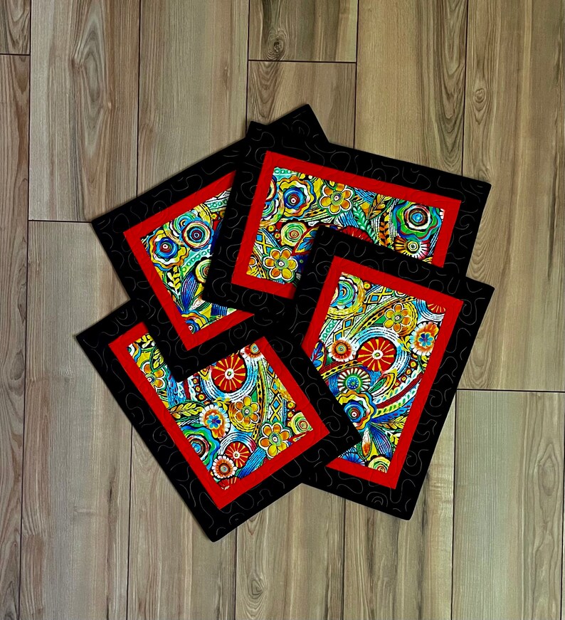 May include: A set of four square coasters with a vibrant floral pattern. Each coaster has a red border, a black patterned frame, and a colorful design featuring flowers and abstract shapes. The coasters are arranged on a wooden surface.