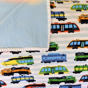 May include: A blue and white fleece blanket with a train pattern. The blanket features a variety of colorful trains, including red, yellow, green, and blue trains. The trains are on a white background with gray stripes.