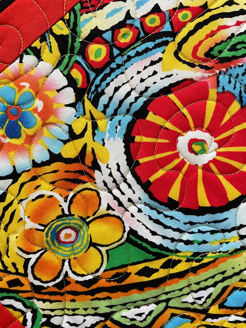 May include: Close-up of a vibrant, quilted fabric with a floral pattern. The design features large, stylized flowers in red, yellow, and orange, set against a black background. The quilting stitches are visible in gold thread, adding texture and detail.