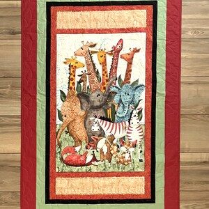 May include: A colorful quilt featuring a whimsical illustration of various animals, including giraffes, elephants, and zebras. The central image is framed by a red border with a leaf pattern, a green and black border, and a light beige border.