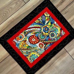 May include: A quilted table runner with a vibrant floral pattern in red, yellow, blue, and green. The central design is framed by a red border and a black outer border with a decorative stitch.