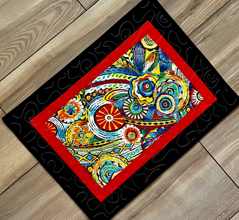 May include: A quilted table runner with a vibrant floral pattern in red, yellow, blue, and green. The central design is framed by a red border and a black outer border with a decorative stitch.