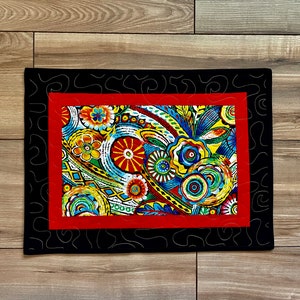 May include: A rectangular, quilted placemat with a vibrant floral design. The central panel features colorful flowers and abstract patterns in red, yellow, blue, and green. It is framed by a red border and a black outer border with a decorative stitch.