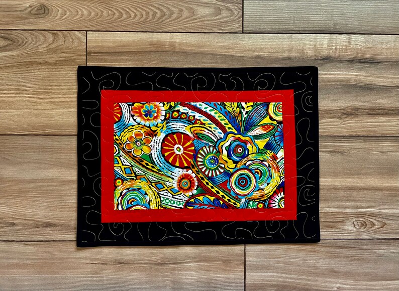 May include: A rectangular, quilted placemat with a vibrant floral design. The central panel features colorful flowers and abstract patterns in red, yellow, blue, and green. It is framed by a red border and a black outer border with a decorative stitch.