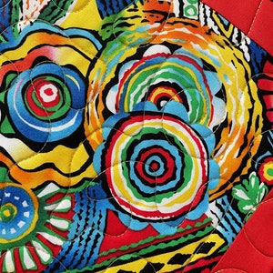 May include: Close-up of a vibrant, colorful quilt with a bold, abstract design. The quilt features a mix of bright colors including red, yellow, blue, and green, with a black border and red binding. The stitching adds texture.