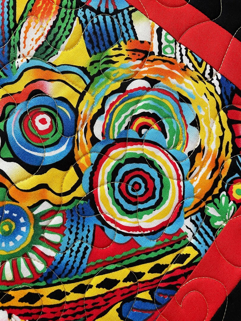 May include: Close-up of a vibrant, colorful quilt with a bold, abstract design. The quilt features a mix of bright colors including red, yellow, blue, and green, with a black border and red binding. The stitching adds texture.