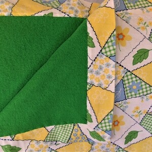 May include: A green fleece fabric square is sewn onto a white fabric background with a patchwork design of yellow, blue, and green squares with floral patterns.