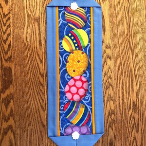 May include: A blue quilted table runner with a row of colorful Easter eggs. The eggs are decorated with stripes, polka dots, and floral patterns.