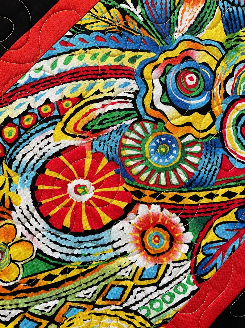 May include: Close-up of a vibrant, quilted fabric with a colorful floral pattern. The design features red, yellow, blue, green, and black elements. The quilting stitches are visible, adding texture to the textile. The edges are framed with a red border.