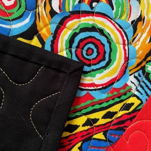 May include: Close-up of a colorful quilt with a black border. The quilt features a vibrant pattern of concentric circles in red, yellow, blue, green, and white. Gold stitching outlines the design and the black border.