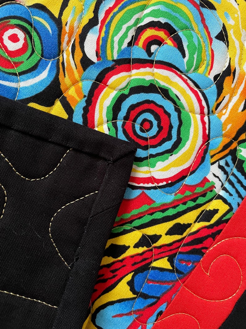 May include: Close-up of a colorful quilt with a black border. The quilt features a vibrant pattern of concentric circles in red, yellow, blue, green, and white. Gold stitching outlines the design and the black border.