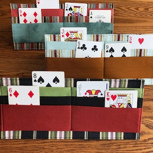 May include: Three fabric card holders with multiple pockets. The top holder is teal, brown, and white striped. The middle holder is brown, white, and blue striped. The bottom holder is red, black, and green striped. Each holder has playing cards inside.
