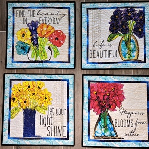 May include: Four square quilted wall hangings with floral designs and inspirational quotes. The first features a bouquet of colorful flowers in a vase with the text "FIND THE beauty IN EVERYDAY". The second features a bouquet of purple flowers in a vase with the text "Life is BEAUTIFUL". The third features a bouquet of yellow flowers in a vase with the text "let your light SHINE". The fourth features a bouquet of red flowers in a vase with the text "Happiness BLOOMS from within".