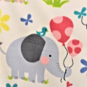 May include: A light blue elephant holding two pink balloons with white polka dots. The elephant is surrounded by green grass, blue and yellow flowers, and a small blue bird.
