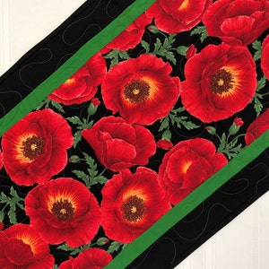 May include: A black table runner with a green border features a repeating pattern of red poppies with yellow centers. The runner is quilted with a black thread.