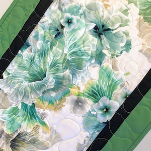 May include: A green and black quilted table runner with a floral pattern featuring green, white, and beige flowers. The runner has a scalloped edge and a black and green border.