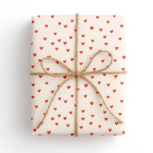 May include: A rectangular gift box wrapped in off-white paper featuring a repeating pattern of small red hearts. The package is tied with a thin, natural brown jute twine string arranged in a cross pattern and finished with a centered bow.