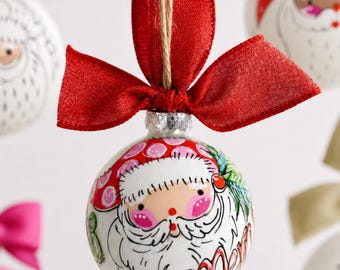 Pode incluir: Um enfeite de Natal esférico de vidro branco com uma ilustração pintada à mão do Pai Natal com bochechas rosadas e barba branca. O enfeite é rematado com um laço de tecido vermelho brilhante e um cordel castanho para pendurar.