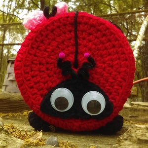 May include: A red crocheted ladybug with black eyes and antennae. The ladybug is sitting on a wooden surface.