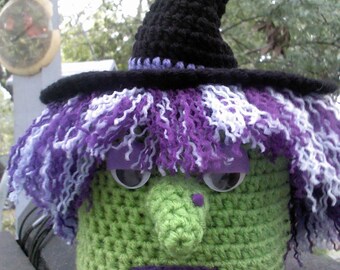 May include: A crocheted witch doll with a black hat, purple and white hair, and a green face. The doll has a large nose and is wearing a purple and white scarf.