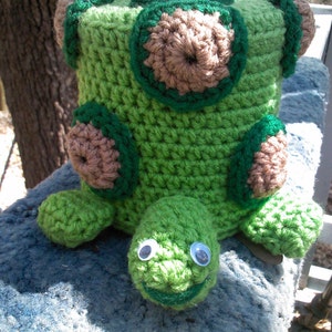 May include: A crocheted green turtle tissue box cover with brown and green accents. The turtle has googly eyes and a smiling mouth.