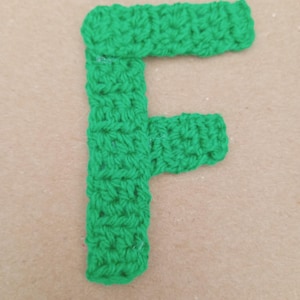 May include: A bright green, crocheted letter "F" against a neutral background. The handmade letter is crafted with a textured stitch, showcasing the detailed craftsmanship. The bold color and simple design make it a striking visual element.