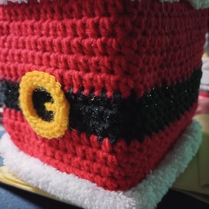 以下が含まれることがあります： 赤いと白い毛糸で編まれたサンタクロースをモチーフにしたティッシュボックスカバー。黒いベルトと黄色のボタンが付いている。