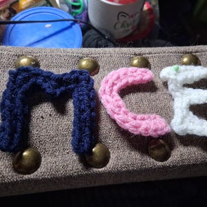 May include: Three individual letters crocheted from yarn, spelling the word MCE. The letter M is dark blue, the letter C is light pink, and the letter E is white. Each letter is constructed with a visible textured stitch pattern.