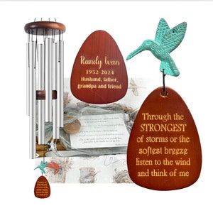 May include: A wooden wind chime with a teal hummingbird ornament and two wooden plaques. The plaques have gold lettering that reads "Randy wan 1952-2024 Husband, father, grandpa and friend" and "Through the STRONGEST of storms or the softest breeze listen to the wind and think of me".