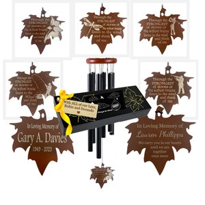 May include: A set of two wind chimes with brown maple leaf-shaped ornaments. The ornaments have engraved messages about strength and remembrance. The chimes are made of black metal tubes and are hung from a wooden frame. The set includes a box with a black background and a white inscription that reads "With all of our love, Robin and Dorando".
