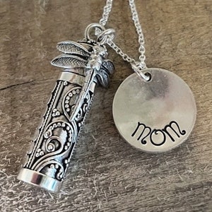 May include: Silver necklace with a small, ornate, cylindrical pendant featuring a dragonfly design. The pendant is attached to a round charm with the word "MOM" engraved on it.