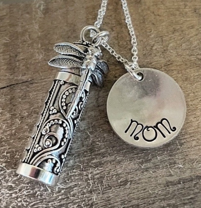May include: Silver necklace with a small, ornate, cylindrical pendant featuring a dragonfly design. The pendant is attached to a round charm with the word "MOM" engraved on it.
