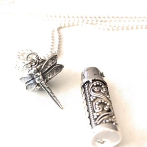 May include: A silver necklace with a dragonfly pendant and a small, cylindrical charm with an ornate design.