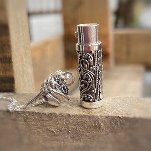 May include: Silver pendant necklace with a small, ornate, cylindrical container and a detailed butterfly charm. The container has a screw-on top and is decorated with intricate floral patterns.