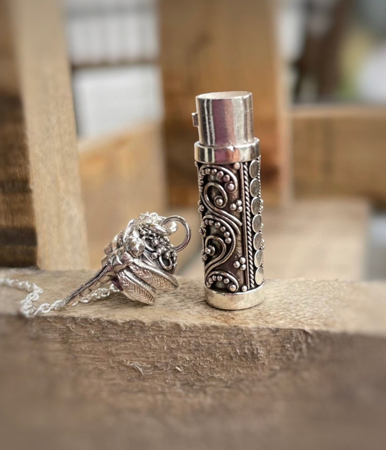 May include: Silver pendant necklace with a small, ornate, cylindrical container and a detailed butterfly charm. The container has a screw-on top and is decorated with intricate floral patterns.