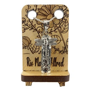 May include: Silver dragonfly cremation necklace with a floral design on a wooden display stand. The stand has the text "Rio Maria Alfred" engraved on it.