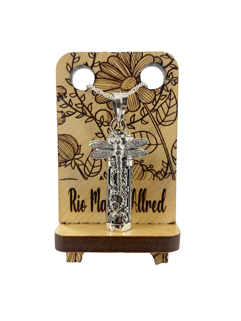 May include: Silver dragonfly cremation necklace with a floral design on a wooden display stand. The stand has the text "Rio Maria Alfred" engraved on it.