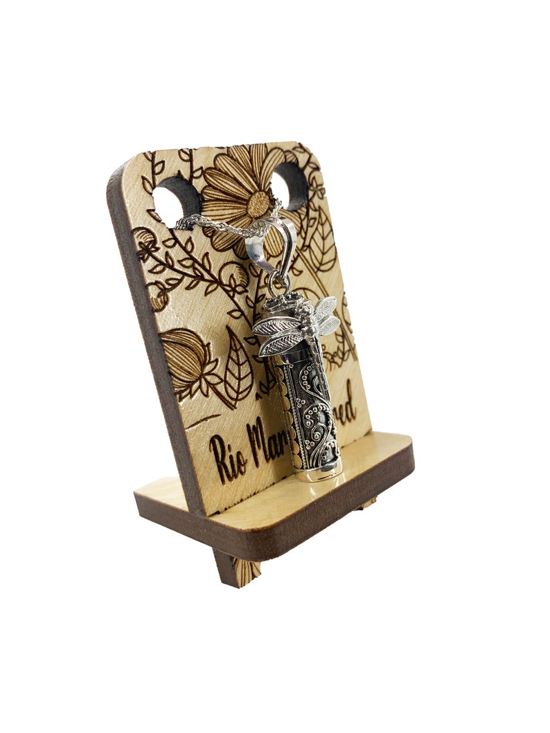 May include: A silver pendant necklace with a dragonfly design and an engraved cylinder charm. The necklace is displayed on a wooden stand with a floral design and the text "Rio Manured".