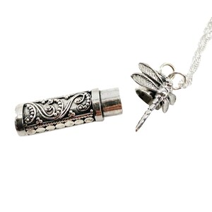 May include: Silver tone necklace with a dragonfly pendant and a small cylindrical container with an ornate design. The container is open and the necklace is attached to the container.