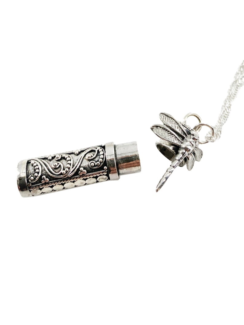 May include: Silver tone necklace with a dragonfly pendant and a small cylindrical container with an ornate design. The container is open and the necklace is attached to the container.