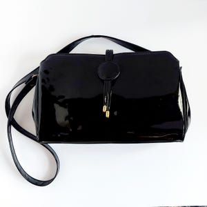 May include: A glossy black handbag with a shoulder strap. The rectangular bag features a flap closure with a circular accent and a drawstring detail with gold-colored tips. The bag is set against a white background.