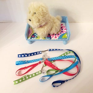 May include: A plush white dog toy lying on a blue and white bed with a floral pattern. The bed has four wooden legs. There are five colourful leashes with white polka dots on a white surface.
