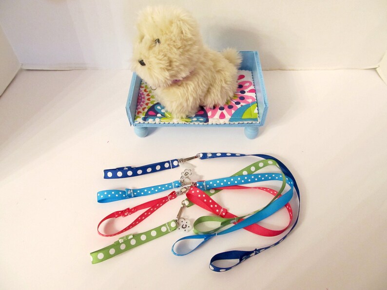 May include: A plush white dog toy lying on a blue and white bed with a floral pattern. The bed has four wooden legs. There are five colourful leashes with white polka dots on a white surface.